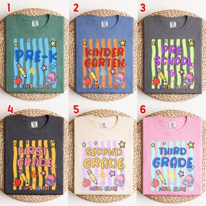 May include: Six crew neck t-shirts in green, blue, dark gray, charcoal, cream, and pink. Each features a graphic with vertical stripes, a pencil, an apple, a backpack, stars, and text including grade levels and the name Mrs. Alice.