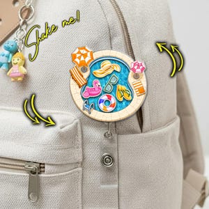 May include: A round, pool-themed badge reel with a retractable cord, attached to a beige backpack. The badge features a pool scene with a flamingo float, sunglasses, and a beach hat. The text "Shake me!" is written in a playful font.