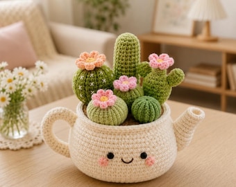 Puede incluir: Una maceta de tetera de ganchillo llena de varias plantas de cactus verdes, cada una coronada con una flor rosa o melocotón. La tetera es de color blanquecino con un diseño de cara sonriente. El arreglo está sobre una mesa de madera.