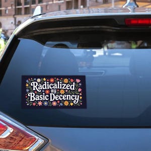 May include: A rectangular sticker on a car window features the text "Radicalized by Basic Decency" in white script. The text is surrounded by a floral border with colorful flowers and green leaves. The sticker is on a dark background.