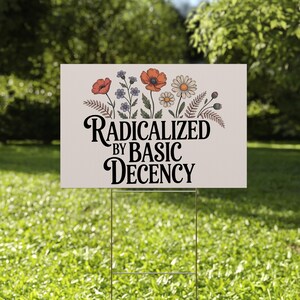 May include: A rectangular yard sign with the text "RADICALIZED BY BASIC DECENCY" in black script. The sign features a floral design with red poppies, daisies, and blue flowers. The sign is displayed on a metal stake in a grassy yard.