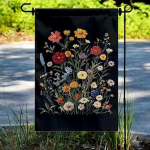May include: A black garden flag featuring a vibrant floral design. The artwork showcases a variety of colorful wildflowers in shades of red, yellow, orange, blue, and white. The flag is displayed on a black metal stand, perfect for outdoor decor.