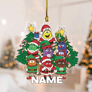 May include: A clear Christmas ornament featuring cartoon characters dressed in Santa and elf costumes. The ornament has a gold string for hanging and includes the word "NAME" at the bottom. The characters are in front of green Christmas trees.