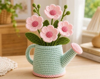 Puede incluir: Un jarrón de ganchillo con forma de regadera en azul claro contiene un ramo de flores rosas con hojas y tallos verdes. Las flores tienen centros rosas. La base del jarrón está adornada con hilo rosa. La regadera está sobre una superficie de madera.