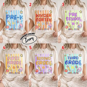 May include: An ivory t-shirt featuring a graphic print with vertical stripes, a pencil, a backpack, stars, and an apple. The text includes grade levels like PRE-K, KINDERGARTEN, PRE SCHOOL, FIRST GRADE, SECOND GRADE, THIRD GRADE, and MRS. ALICE.