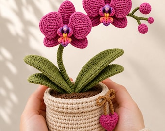 Puede incluir: Una planta de orquídea tejida con flores magenta vibrantes, hojas verdes y una maceta beige. La maceta tiene un interior marrón y un pequeño dije de corazón magenta. La planta hecha a mano es un artículo decorativo.