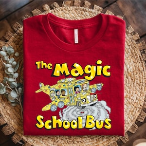 May include: A red t-shirt featuring a yellow school bus with wings, the text "The Magic School Bus," and cartoon characters. The bus is emitting a cloud of smoke. The shirt is folded on a woven surface.