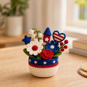 May include: A crocheted floral arrangement in a white, red, and blue pot. The arrangement includes a red rose, white daisy, blue flowers, a blue star, a red and blue heart, and a red, white, and blue firework. The pot has a red and blue striped design.