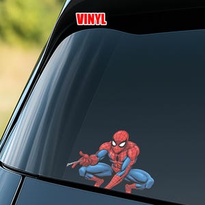 May include: A red and blue Spider-Man sticker is displayed on a car window. The sticker features the superhero in a crouching pose, with the word "VINYL" in red block letters at the top. The background is a blurred view of a green landscape.