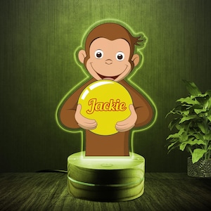 May include: A brown monkey-shaped night light holding a yellow ball with the name "Jackie" in red script. The night light sits on a wooden base and emits a green glow. The background is a textured green.
