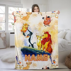 May include: A soft, plush blanket featuring a watercolor-style design of characters from the Lion King movie. The blanket features Simba, Nala, Mufasa, Pumbaa, and Timon. The blanket is white with yellow, orange, and brown accents. The text "NAME" is printed in gold letters on the blanket.