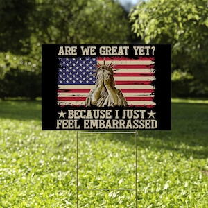 May include: A black and white yard sign with an American flag and the Statue of Liberty. The sign reads "Are we great yet? Because I just feel embarrassed."