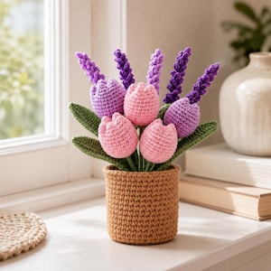May include: A handcrafted crochet flower arrangement featuring pink tulips, purple lavender, and green leaves in a brown crochet pot. The arrangement is displayed on a white surface near a window.