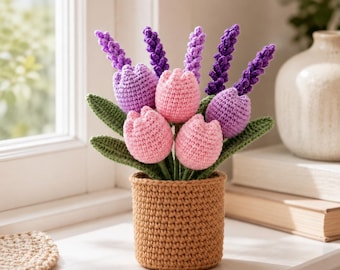 Puede incluir: Un arreglo floral de ganchillo hecho a mano con tulipanes rosas, lavanda morada y hojas verdes en una maceta de ganchillo marrón. El arreglo se exhibe sobre una superficie blanca cerca de una ventana.