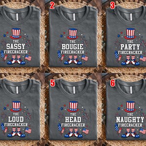 May include: A collection of six dark gray t-shirts, each featuring a graphic of a red, white, and blue top hat, fireworks, a rocket, a small American flag, and shoes. The text reads THE SASSY, BOUGIE, PARTY, LOUD, HEAD, or NAUGHTY FIRECRACKER.