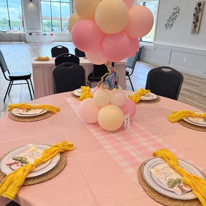 Könnte beinhalten: Ein Tischmittelstück bestehend aus einem Bündel pastellrosa und cremefarbener Luftballons mit einem Eeyore-Ausschnitt. Der Tisch ist mit einer rosa Tischdecke, einem rosa-weiß karierten Läufer und Gedecken mit gelben Servietten und Platzsets gedeckt.