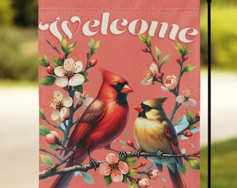 May include: A garden flag with a coral background features a pair of cardinals perched on a flowering branch. The word "welcome" is written in white script across the top. The flag is displayed on a black metal stand.