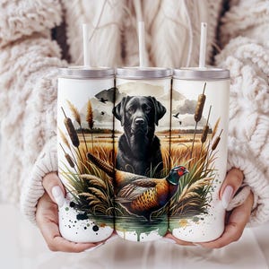 Op de afbeelding: Drie witte geïsoleerde bekers met zilveren deksels en rietjes. Elke beker heeft een aquarel illustratie van een zwarte Labrador, fazant en moerasgrassen. Het ontwerp omvat een landschap met lisdodden en vliegende vogels.