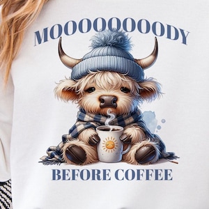 May include: A white graphic sweatshirt featuring a cartoon Highland cow wearing a blue knit hat and scarf, holding a mug with a smiling sun. The text "MOOOOOOODY" is arched above the cow, and "BEFORE COFFEE" is below.