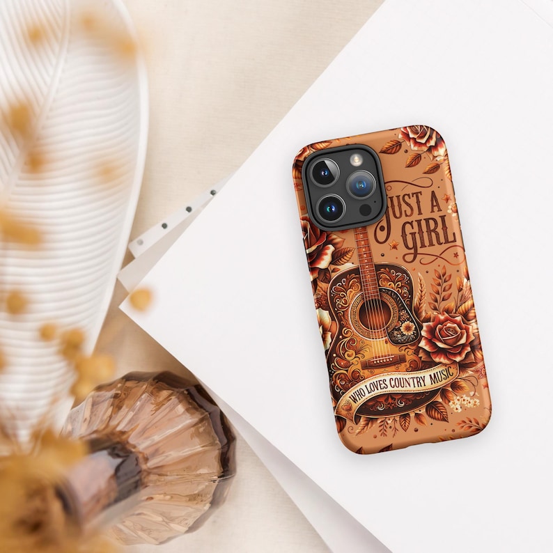 May include: A phone case with a brown background featuring a detailed illustration of an acoustic guitar, roses, and the text "Just a Girl Who Loves Country Music." The case has a glossy finish and a camera cutout.