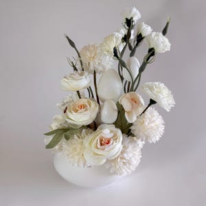May include: A white ceramic vase filled with a bouquet of white and cream artificial flowers. The flowers include carnations, roses, and chrysanthemums.