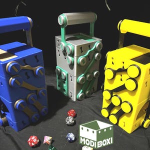 May include: Three colourful, stackable dice storage boxes with handles. The boxes are blue, silver, and yellow. Each box has a different pattern of circular cutouts. The boxes are sitting on a black surface with dice scattered around them. The text "MODI BOXI" is visible in the lower right corner of the image.