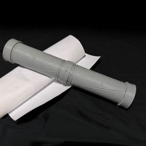 May include: A gray, cylindrical object with a textured surface and a decorative band. The object is lying on a white piece of paper.