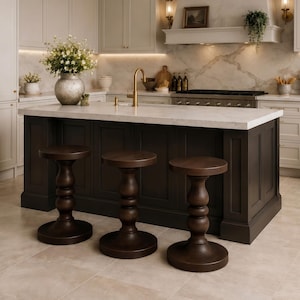 Puede incluir: Isla de cocina con encimera de mármol y armarios de color marrón oscuro. Tres taburetes de bar de madera con asientos redondos y patas torneadas están frente a la isla. Un jarrón con flores está sobre la encimera.
