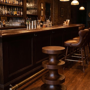 May include: A dark brown wooden stool with a stacked, cylindrical, hourglass-inspired design featuring three circular tiers. The stool is positioned in front of a long, dark wood bar counter with a brass foot rail and brown leather bar stools.