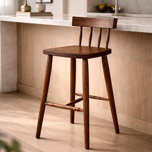 Könnte beinhalten: Ein Barhocker aus dunkelbraunem Holz. Er hat eine quadratische Sitzfläche, eine Rückenlehne mit vier vertikalen Sprossen und vier abgewinkelten Beinen mit einer Fußstütze. Der Hocker steht in einer Küche. Höhe ca. 76 cm.