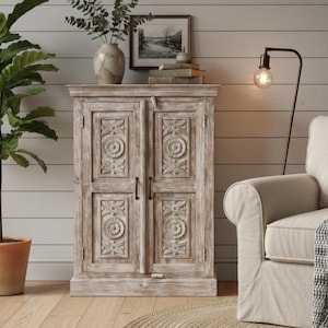May include: A light-coloured wooden cabinet with carved floral designs on the double doors. The cabinet has a distressed finish and sits on a wooden floor. A vase with greenery and a framed picture are on top.