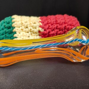 May include: A glass pipe with orange, yellow, and blue swirls. The pipe is lying on a colorful crocheted fabric.