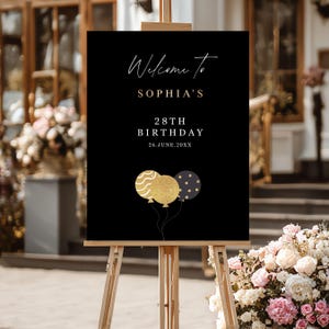May include: A black welcome sign on a wooden easel. The sign reads "Welcome to SOPHIA'S 28TH BIRTHDAY 26.JUNE.20XX" in gold and white script. Three gold and navy blue balloons are at the bottom of the sign.