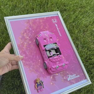 May include: A framed pink Lego car model with the text "The Fast and the Furious". The car is a convertible with silver wheels and a small Lego figure. The background is pink with a Honda logo.