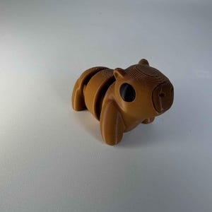 Op de afbeelding: Een bruin, gearticuleerd capybarafiguur. Het speelgoed is gemaakt van gelaagd materiaal, met zichtbare lijnen en een gladde afwerking. De capybara heeft grote, zwarte ogen en bevindt zich in rustpositie. Het beeldje is een uniek decoratief item.