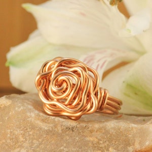 May include: A copper wire ring shaped like a rose, resting on a light-colored stone. The rose is made of intricately coiled wire, with a detailed center. The ring is set against a blurred background of a white flower.