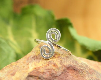 May include: A silver-colored ring with a double spiral design. The ring is set on a brown rock with a green leaf in the background. The ring's design features two swirling spirals connected by a thin band.