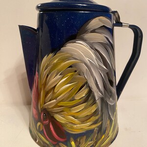 May include: A blue enamel coffee pot with a rooster painted on the side. The rooster is yellow, white, and red with a black eye. The pot has a black handle and a lid.