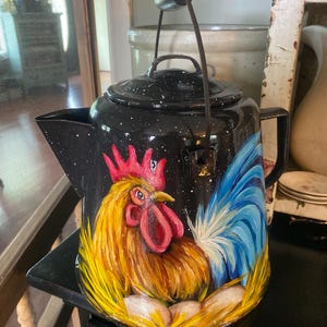 May include: A black speckled enamel coffee pot featuring a hand-painted rooster with a bright red comb, yellow neck feathers, and blue tail feathers, sitting in a yellow nest with white eggs. The pot has a metal handle and a matching lid.