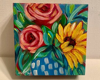 May include: A small, colorful painting on wood featuring a sunflower and two pink roses. The flowers are painted in a vibrant style with bright yellow, pink, and red hues. The background is a teal blue with white polka dots. The painting is signed "2024 SLP" in the lower left corner.