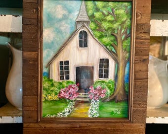 May include: A painting of a white church with a steeple, surrounded by green trees and pink flowers. The church has a wooden door and windows. The painting is framed in a rustic wooden frame. The text "Cottage Design Studio" is below the frame.