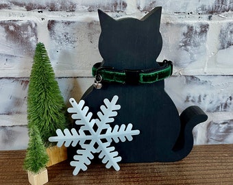 May include: A black cat-shaped wooden decoration with a green velvet cat collar and a small silver bell. A white snowflake and two small green Christmas trees are also present. The background is a white brick wall.