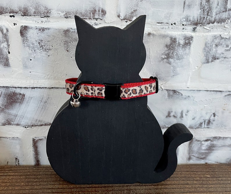 May include: A black cat-shaped wooden decoration with a red and leopard print cat collar. The collar has a small silver bell attached. The background is a white brick wall.