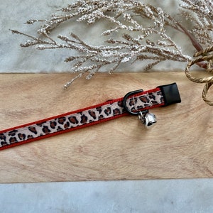 May include: A red and leopard print cat collar with a silver bell. The collar has a black buckle and a tan and brown leopard print pattern. The collar is on a wooden surface.