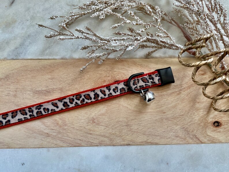May include: A red and leopard print cat collar with a silver bell. The collar has a black buckle and a tan and brown leopard print pattern. The collar is on a wooden surface.