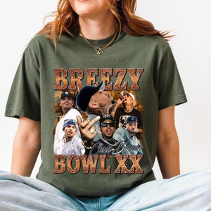A woman wearing a green t-shirt with a graphic design featuring a group of people and the text "BREZY BOWL XX" is sitting on the floor.