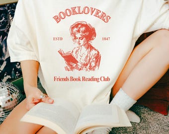 May include: Cream-colored t-shirt with a red graphic that reads "BOOKLOVERS" above an illustration of a woman reading. Below the illustration are the words "Friends Book Reading Club" and the year "1847".