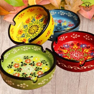 以下が含まれることがあります： 花柄のカラフルなセラミック灰皿4つ。 灰皿は黄色、青、赤、緑で、それぞれ黒い縁とユニークな花と葉の模様があります。 木の表面に置かれています。