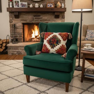 May include: A cozy living room scene featuring a dark green armchair with a decorative, square pillow. The pillow has a white background with a colorful, geometric design and tassels. A fireplace with a burning fire is in the background.