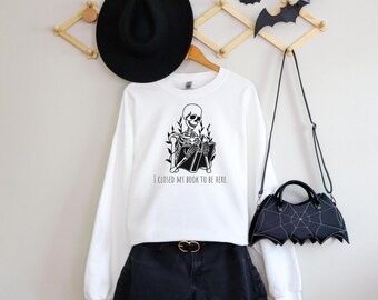 May include: White sweatshirt with a black skeleton reading a book and the text "I closed my book to be here". The sweatshirt is paired with black denim shorts and a black bat-shaped handbag.